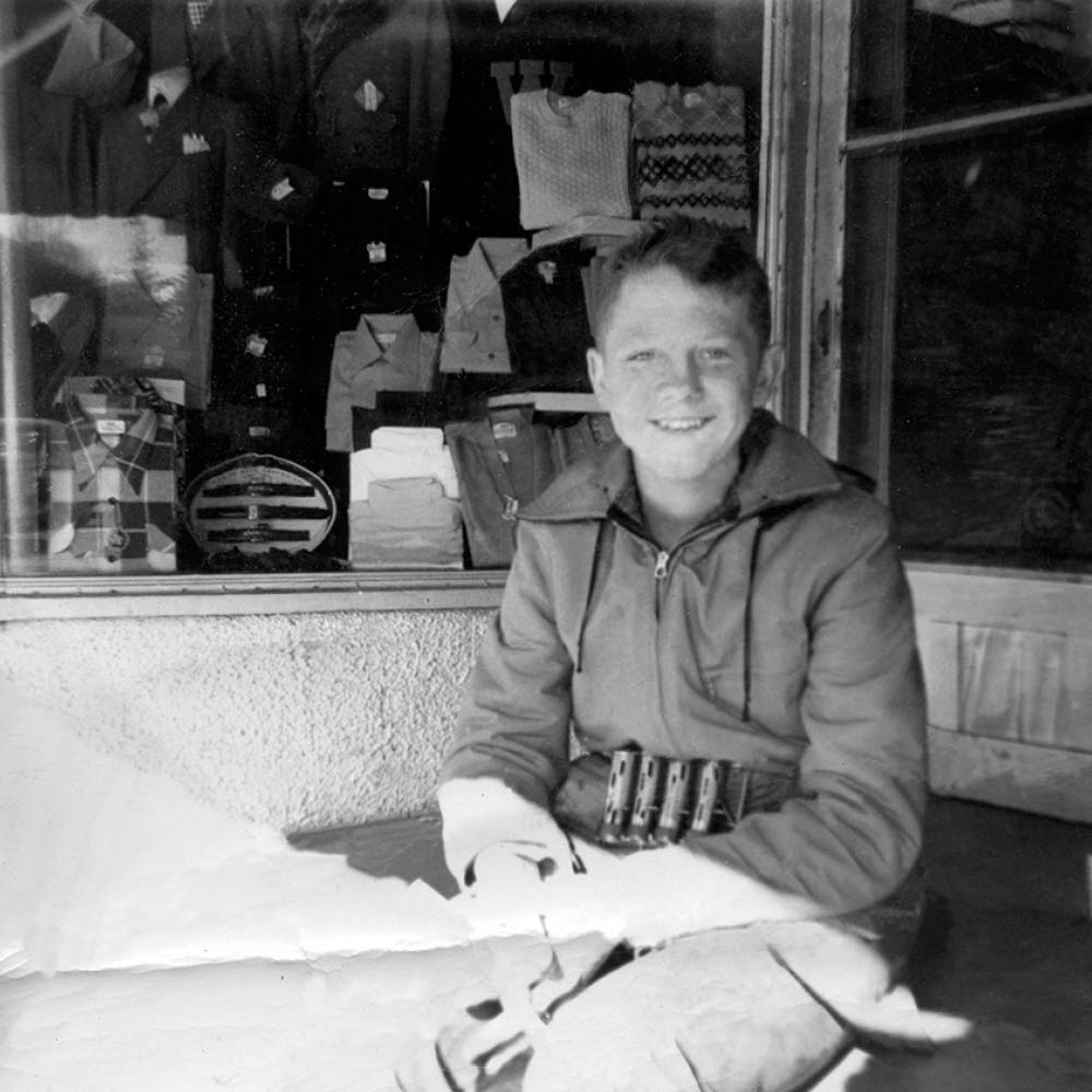 Black and white photo of Alfred Ludeking at age 12 sitting on a shop stoop selling newspapers
